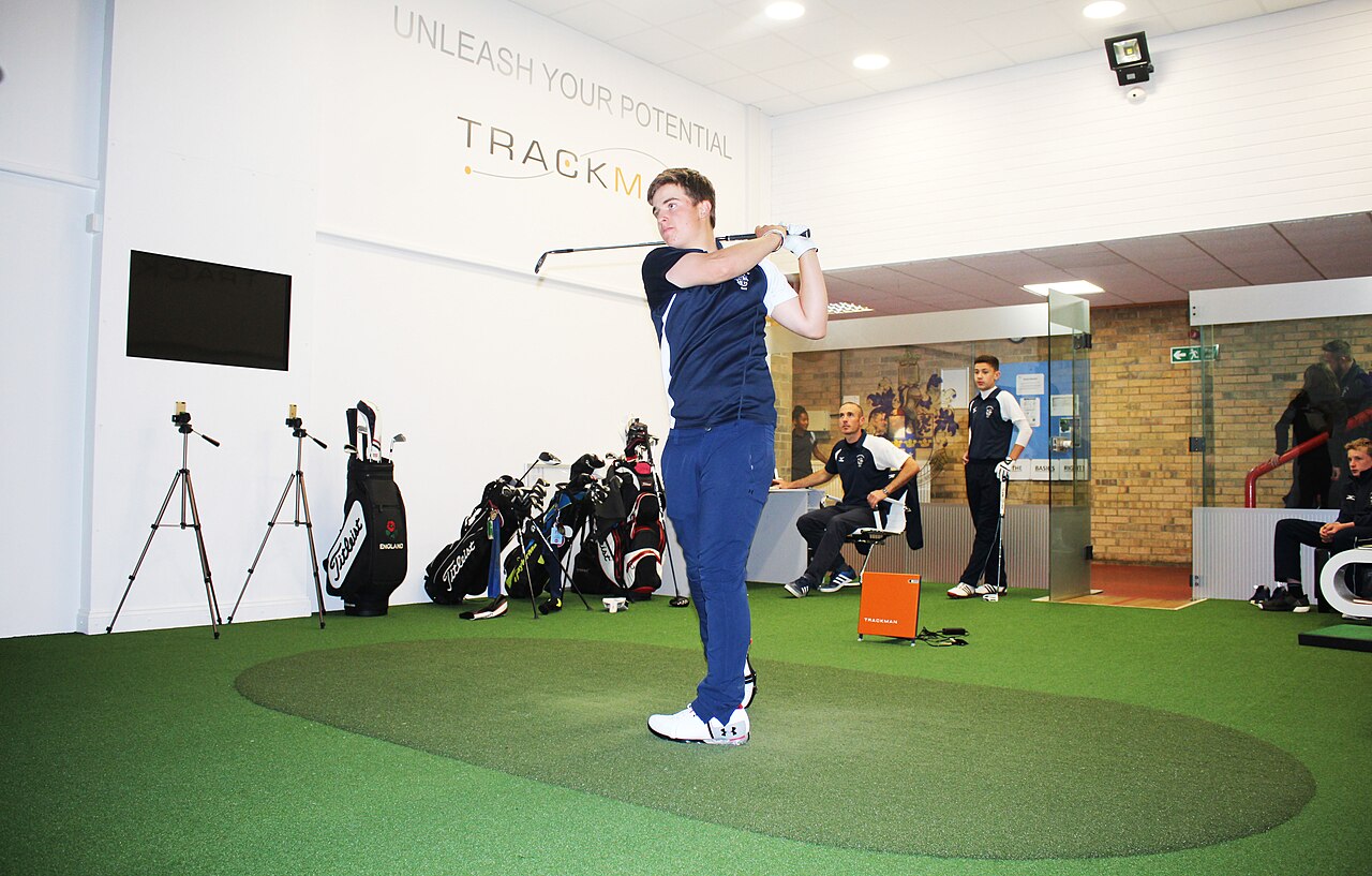 A TrackMan launch monitor unit in an indoor golf studio, with a golfer at the top of his backswing